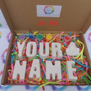 May include: A craft kit in a cardboard box, featuring the words "YOUR NAME" in large, unpainted letters. The box includes paint pots in various colors, a small figurine, and colorful paper shreds for decoration. The text "Set: N8" is visible.