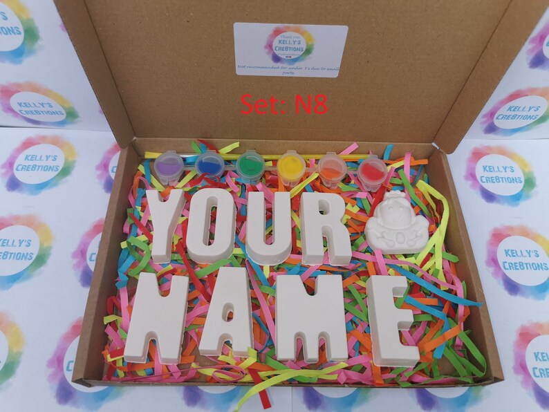 May include: A craft kit in a cardboard box, featuring the words "YOUR NAME" in large, unpainted letters. The box includes paint pots in various colors, a small figurine, and colorful paper shreds for decoration. The text "Set: N8" is visible.