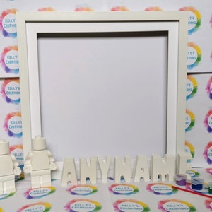 May include: A white picture frame with the word "ANYNAME" in white block letters. Two white Lego figures stand near the frame. Small paint pots and a paintbrush are also visible.
