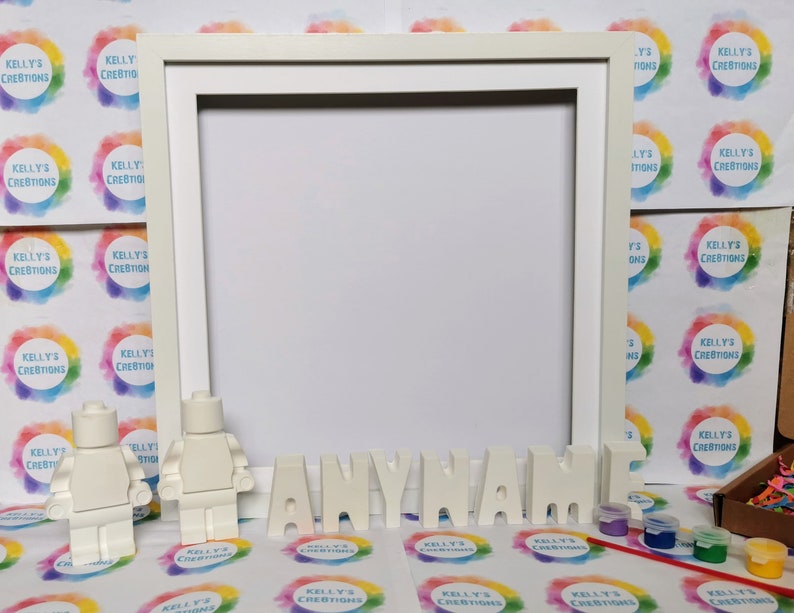 May include: A white picture frame with the word "ANYNAME" in white block letters. Two white Lego figures stand near the frame. Small paint pots and a paintbrush are also visible.
