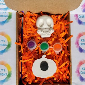 May include: A Halloween-themed craft kit in a cardboard box. The kit includes a white skull and pumpkin, three paint pots, and a paintbrush. The box is filled with orange paper shreds. The text "KELLY'S CREATIONS" is visible on the box.