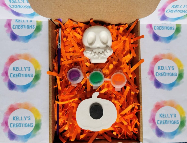 May include: A Halloween-themed craft kit in a cardboard box. The kit includes a white skull and pumpkin, three paint pots, and a paintbrush. The box is filled with orange paper shreds. The text "KELLY'S CREATIONS" is visible on the box.