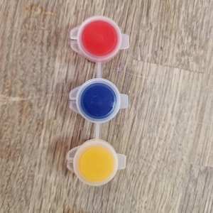 May include: Three connected paint pots, each filled with a different color: red, blue, and yellow. The pots are made of white plastic and are arranged vertically against a wooden background.