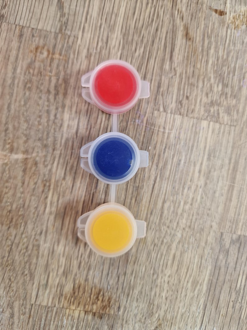 May include: Three connected paint pots, each filled with a different color: red, blue, and yellow. The pots are made of white plastic and are arranged vertically against a wooden background.