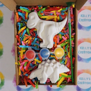 Op de afbeelding: Een knutselpakket in een kartonnen doos, met twee witte dinosaurusfiguren, drie verfpotjes met rode, blauwe en gele verf en een groene penseel. De doos is gevuld met kleurrijk papier.
