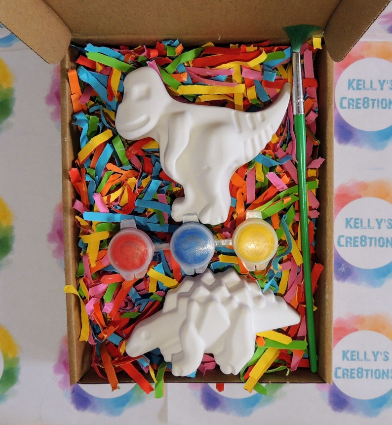 Op de afbeelding: Een knutselpakket in een kartonnen doos, met twee witte dinosaurusfiguren, drie verfpotjes met rode, blauwe en gele verf en een groene penseel. De doos is gevuld met kleurrijk papier.