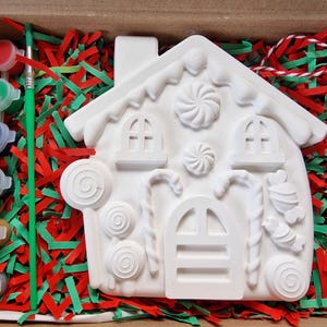 May include: A white ceramic gingerbread house ornament with candy cane details, lollipops, and a candy swirl design. The ornament is ready to be painted with the included set of paint pots.