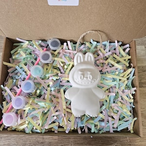 Peut inclure: Un kit de bricolage dans une bo&icirc;te en carton, comprenant une figurine blanche en forme de lapin avec un visage souriant. Le kit comprend de petits pots de peinture de diff&eacute;rentes couleurs, un pinceau et du papier d&eacute;chiquet&eacute; color&eacute;.