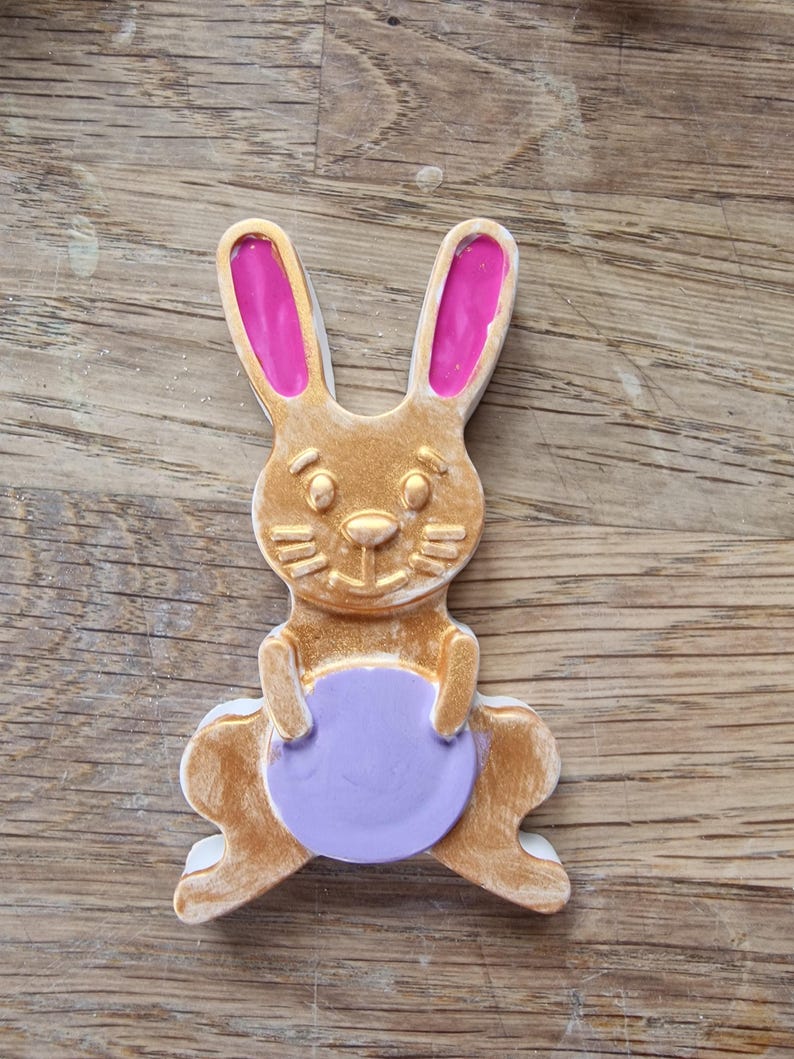 May include: A gold bunny rabbit figurine with pink ears holding a purple egg.