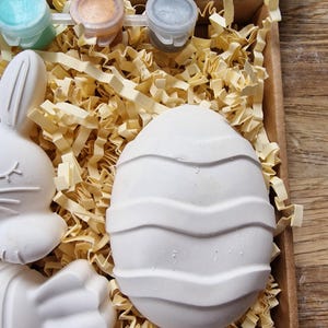 May include: A craft kit with three small paint pots, a paintbrush, and three ceramic figurines. The figurines are a bunny, a paw print, and an egg with a wavy design. The kit is packaged in a brown cardboard box with shredded paper.