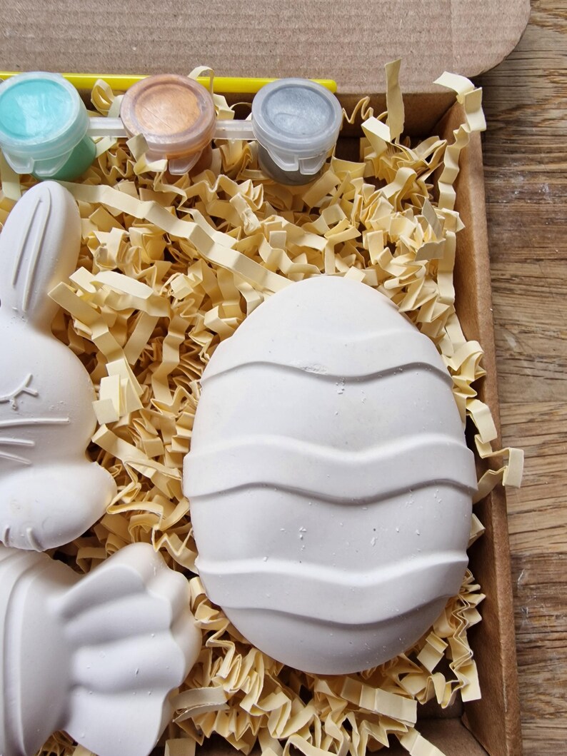 May include: A craft kit with three small paint pots, a paintbrush, and three ceramic figurines. The figurines are a bunny, a paw print, and an egg with a wavy design. The kit is packaged in a brown cardboard box with shredded paper.