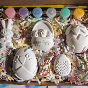 May include: A set of six white ceramic Easter-themed figurines in a box with colorful shredded paper. The figurines include a bunny, a basket with the words "Happy Easter", and four eggs with different designs. The set also includes six small bottles of paint and a paintbrush.
