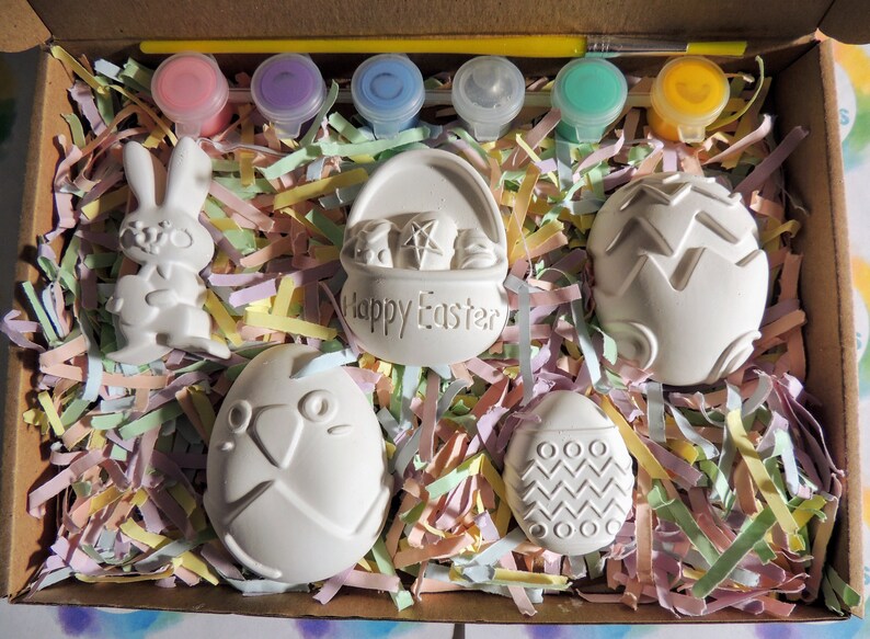 May include: A set of six white ceramic Easter-themed figurines in a box with colorful shredded paper. The figurines include a bunny, a basket with the words "Happy Easter", and four eggs with different designs. The set also includes six small bottles of paint and a paintbrush.