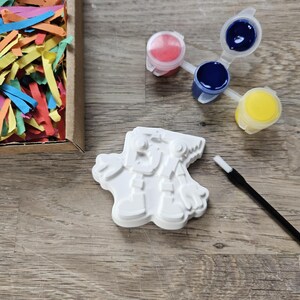 May include: A white snowman plaster figure, paint pots with red, blue, and yellow paint, and a paintbrush are arranged on a wooden surface. A cardboard box filled with colourful paper strips is also present, suggesting a craft or painting kit.