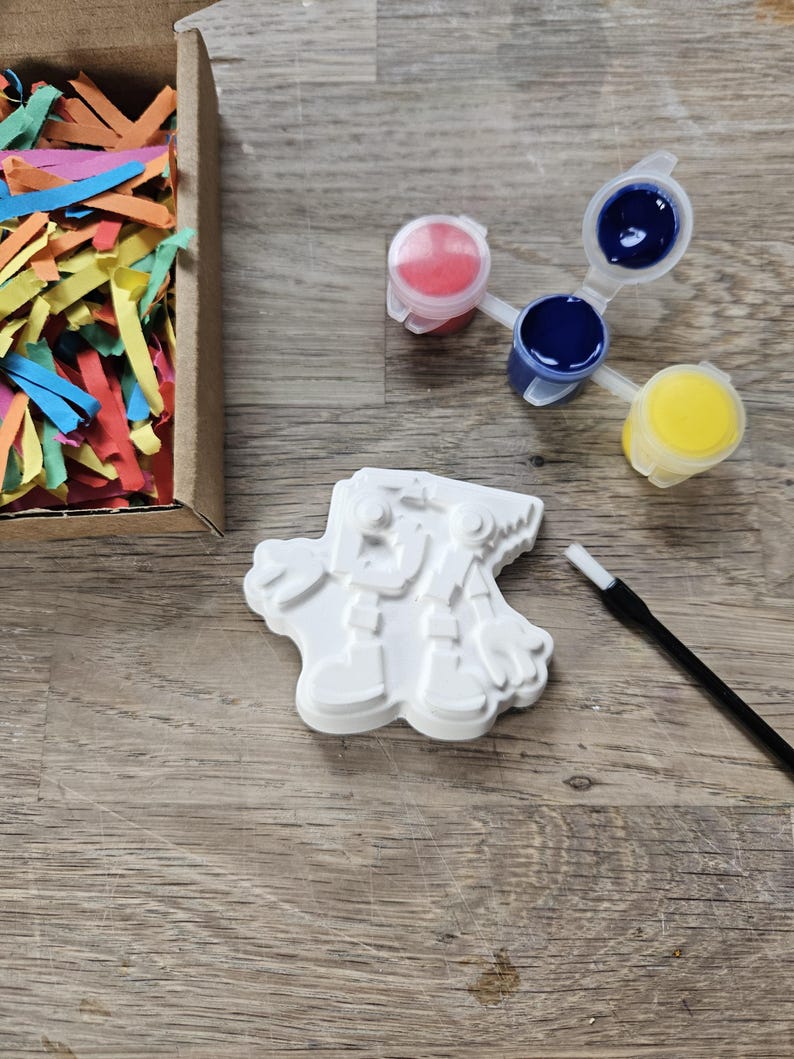 May include: A white snowman plaster figure, paint pots with red, blue, and yellow paint, and a paintbrush are arranged on a wooden surface. A cardboard box filled with colourful paper strips is also present, suggesting a craft or painting kit.