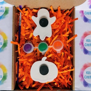 May include: A Halloween-themed craft kit in a cardboard box. The kit includes a ghost and pumpkin-shaped magnet, paint pots with purple, green, and orange paint, and a paintbrush. The box is filled with orange paper shreds.