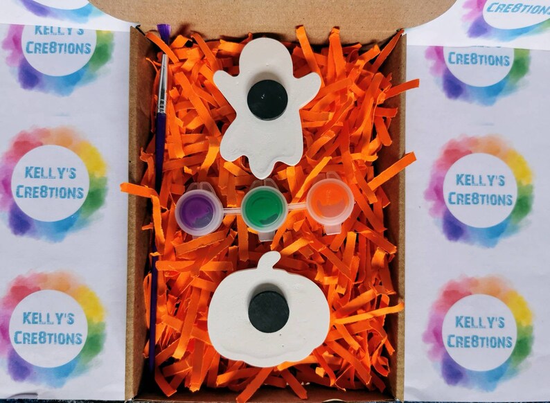 May include: A Halloween-themed craft kit in a cardboard box. The kit includes a ghost and pumpkin-shaped magnet, paint pots with purple, green, and orange paint, and a paintbrush. The box is filled with orange paper shreds.
