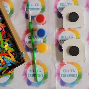 May include: A craft kit with three paint pots, a paintbrush, and two white ceramic shapes. The paint pots are red, blue, and yellow. The ceramic shapes are shaped like a bus and have a black circle in the middle. The kit is labeled "Kelly's Creations".