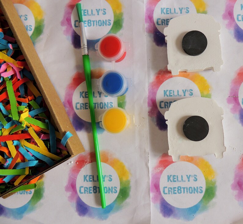 May include: A craft kit with three paint pots, a paintbrush, and two white ceramic shapes. The paint pots are red, blue, and yellow. The ceramic shapes are shaped like a bus and have a black circle in the middle. The kit is labeled "Kelly's Creations".