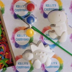 May include: A craft kit with two white ceramic dinosaur figurines, a paintbrush, and three small paint pots with red, blue, and yellow paint. The paint pots are labeled "Kelly's Creations".