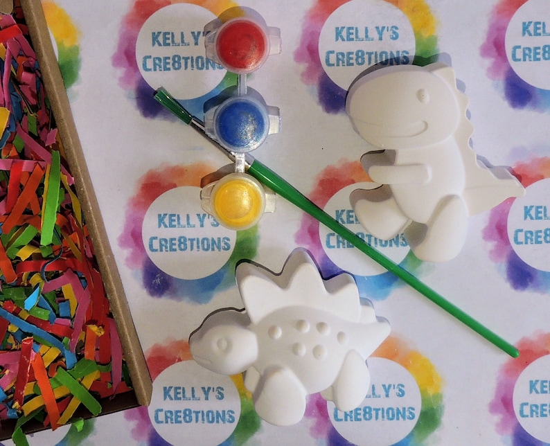 May include: A craft kit with two white ceramic dinosaur figurines, a paintbrush, and three small paint pots with red, blue, and yellow paint. The paint pots are labeled "Kelly's Creations".