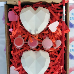 May include: A craft kit with two white geometric heart-shaped ceramic ornaments, three small paint pots, a paintbrush, and pink paper hearts in a cardboard box filled with red shredded paper.