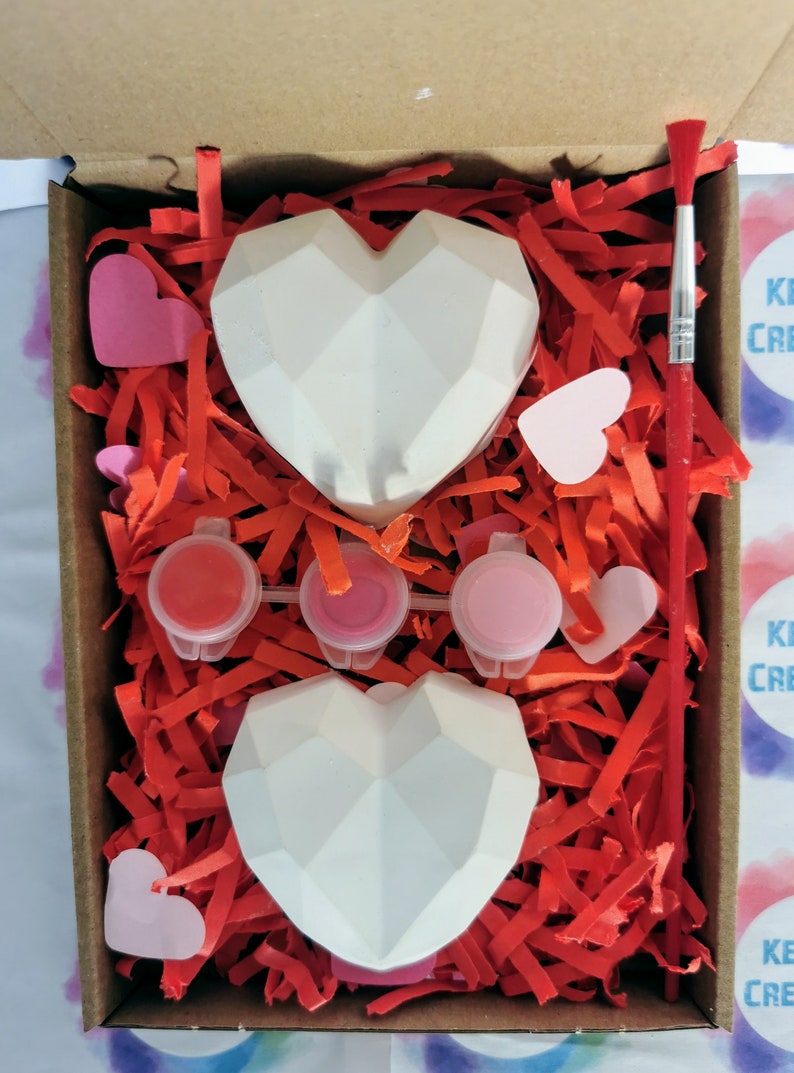 May include: A craft kit with two white geometric heart-shaped ceramic ornaments, three small paint pots, a paintbrush, and pink paper hearts in a cardboard box filled with red shredded paper.