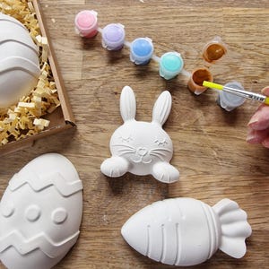 May include: A set of Easter-themed ceramic painting projects. The set includes a bunny, an egg, and a carrot. The ceramic pieces are white and ready to be painted. A set of paint pots with different colors is also included.