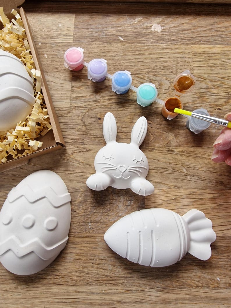 May include: A set of Easter-themed ceramic painting projects. The set includes a bunny, an egg, and a carrot. The ceramic pieces are white and ready to be painted. A set of paint pots with different colors is also included.