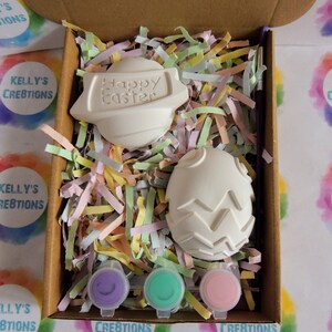 May include: A craft kit containing two white ceramic Easter eggs, one with the words "Happy Easter" and the other with a textured design. The kit includes three small pots of paint in purple, green and pink.