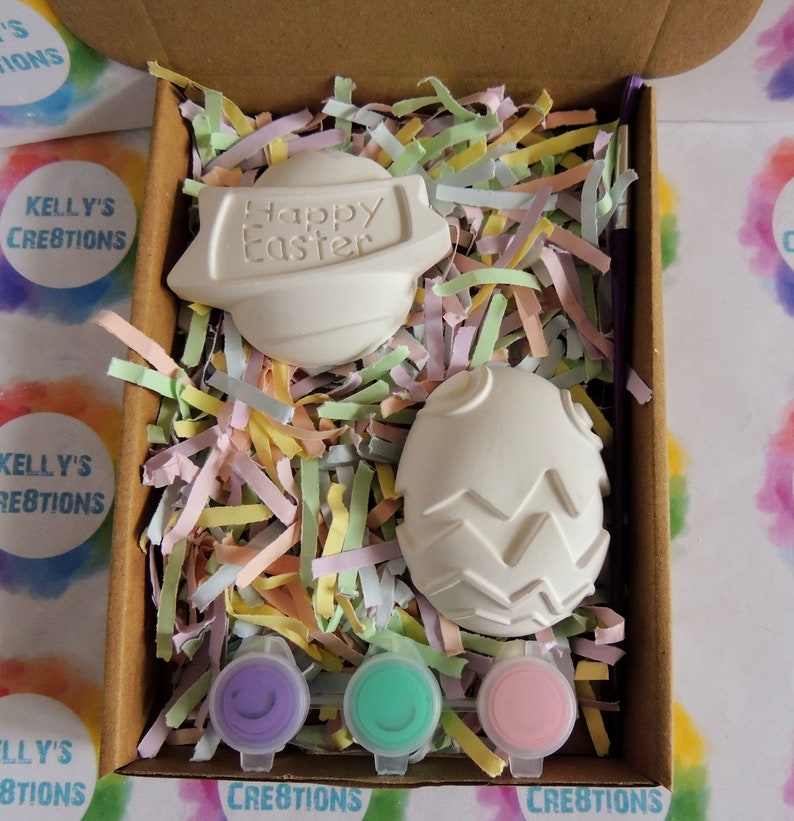 May include: A craft kit containing two white ceramic Easter eggs, one with the words "Happy Easter" and the other with a textured design. The kit includes three small pots of paint in purple, green and pink.