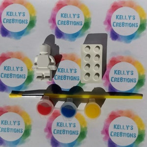 Puede incluir: Una figurita blanca de pl&aacute;stico de una persona, un molde para hacer un ladrillo y tres peque&ntilde;os botes de pintura roja, azul y amarilla. Los botes de pintura est&aacute;n etiquetados con "Kelly's Creations".