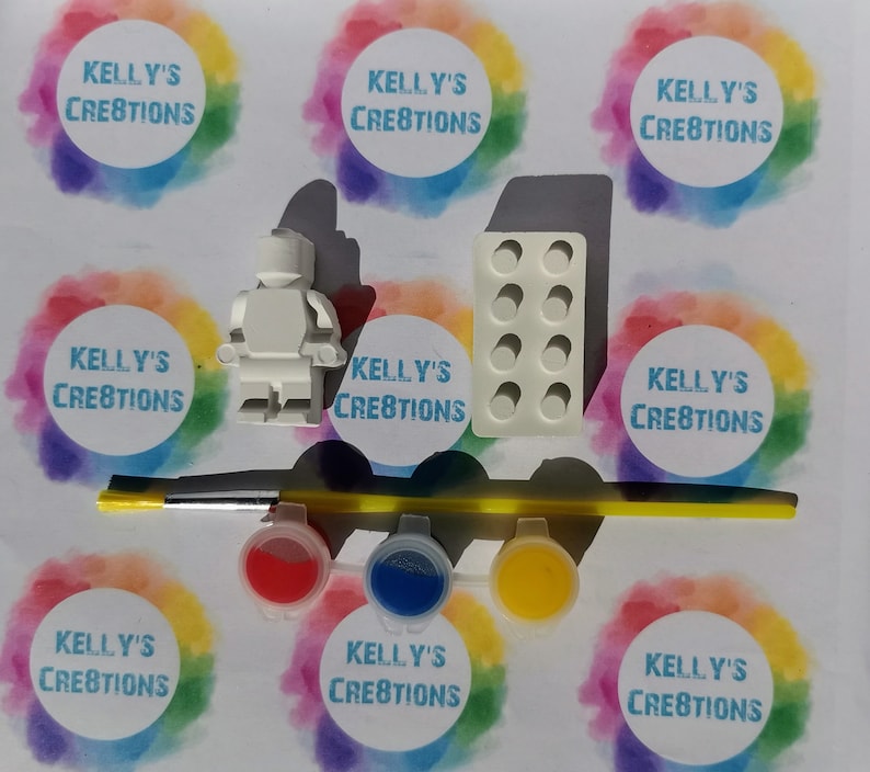 Puede incluir: Una figurita blanca de pl&aacute;stico de una persona, un molde para hacer un ladrillo y tres peque&ntilde;os botes de pintura roja, azul y amarilla. Los botes de pintura est&aacute;n etiquetados con "Kelly's Creations".