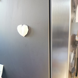 May include: A white, geometric heart-shaped magnet is attached to a stainless steel refrigerator door.