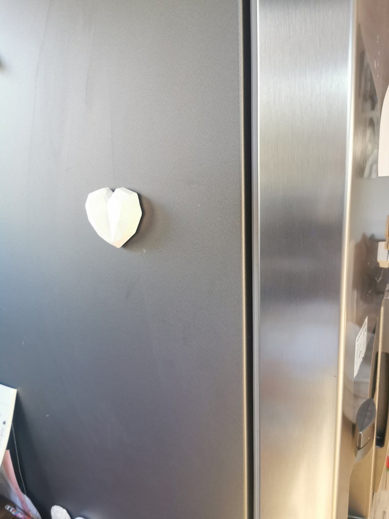 May include: A white, geometric heart-shaped magnet is attached to a stainless steel refrigerator door.