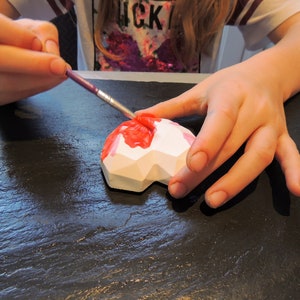 May include: A person paints a white geometric heart-shaped object with pink paint using a paintbrush. The object is on a black surface.