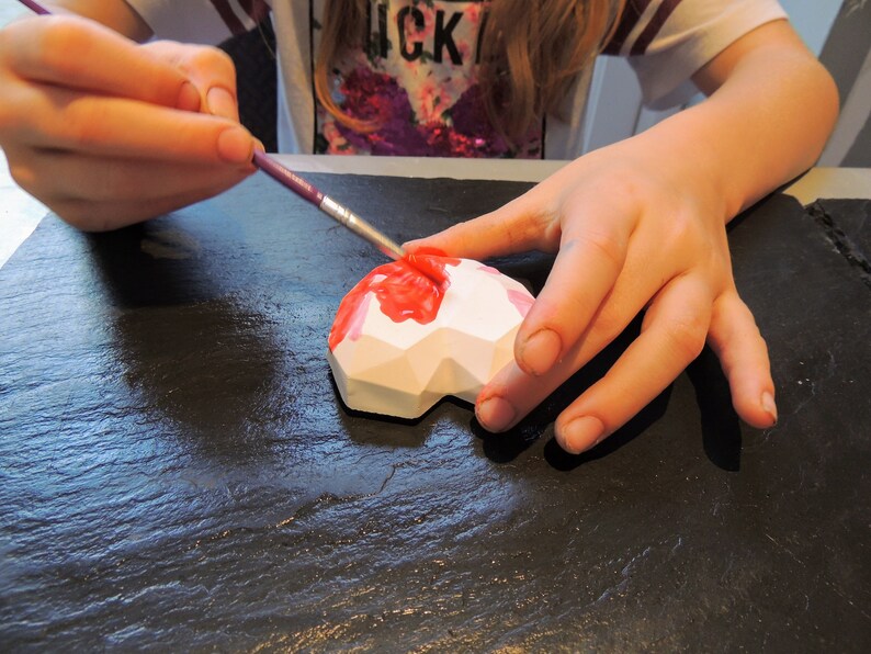 May include: A person paints a white geometric heart-shaped object with pink paint using a paintbrush. The object is on a black surface.