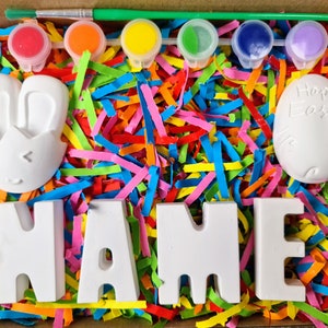 May include: A craft kit with white ceramic bunny, egg, and letters spelling "NAME" in a box filled with colorful shredded paper. The kit includes five small paint pots with red, orange, yellow, green, and purple paint.
