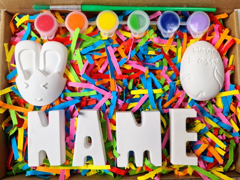 May include: A craft kit with white ceramic bunny, egg, and letters spelling "NAME" in a box filled with colorful shredded paper. The kit includes five small paint pots with red, orange, yellow, green, and purple paint.