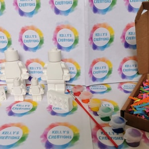 May include: A family of three white blank Lego minifigures, two small and one large, with a white Lego brick, a paintbrush, and six small jars of paint. The paint colors are red, yellow, orange, green, blue, and purple. The items are on a white surface with a colorful background.