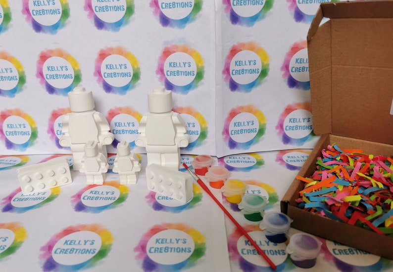 May include: A family of three white blank Lego minifigures, two small and one large, with a white Lego brick, a paintbrush, and six small jars of paint. The paint colors are red, yellow, orange, green, blue, and purple. The items are on a white surface with a colorful background.