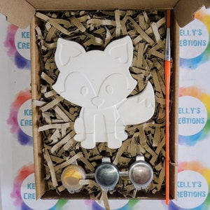 May include: A paint-your-own ceramic fox figurine kit. The kit includes a white ceramic fox, three small paint pots, and a paintbrush. The fox is sitting on a bed of brown packing peanuts.
