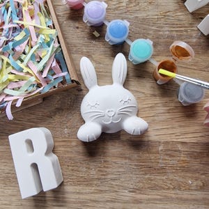 May include: A white ceramic bunny figurine with closed eyes and a small nose, ready to be painted.  The bunny is surrounded by paint pots in various colors, including pink, purple, blue, turquoise, brown, and gray.