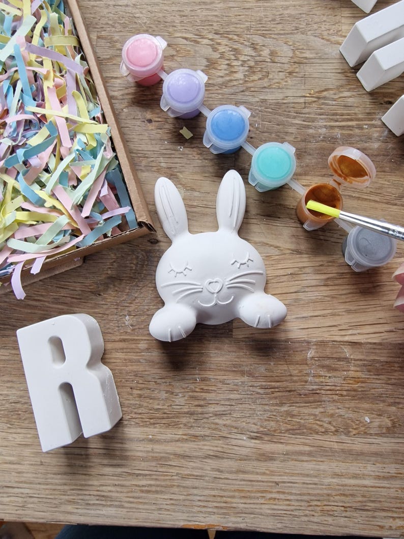 May include: A white ceramic bunny figurine with closed eyes and a small nose, ready to be painted.  The bunny is surrounded by paint pots in various colors, including pink, purple, blue, turquoise, brown, and gray.