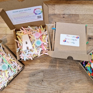 May include: Four small cardboard boxes with lids open, each containing a white plaster letter, paint pots, and a paintbrush. The letters spell out "B.A.S.". The boxes are filled with colourful shredded paper. One box has a thank you tag that says "Thank you for coming to my party."