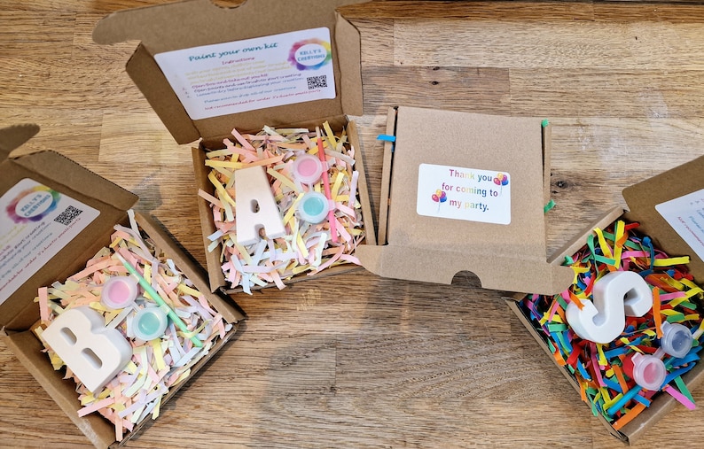 May include: Four small cardboard boxes with lids open, each containing a white plaster letter, paint pots, and a paintbrush. The letters spell out "B.A.S.". The boxes are filled with colourful shredded paper. One box has a thank you tag that says "Thank you for coming to my party."