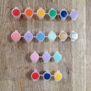 May include: A collection of small, plastic paint pots filled with various colors, including red, orange, yellow, green, blue, and purple. The paint pots are arranged in rows on a wooden surface, offering a vibrant palette for art projects.