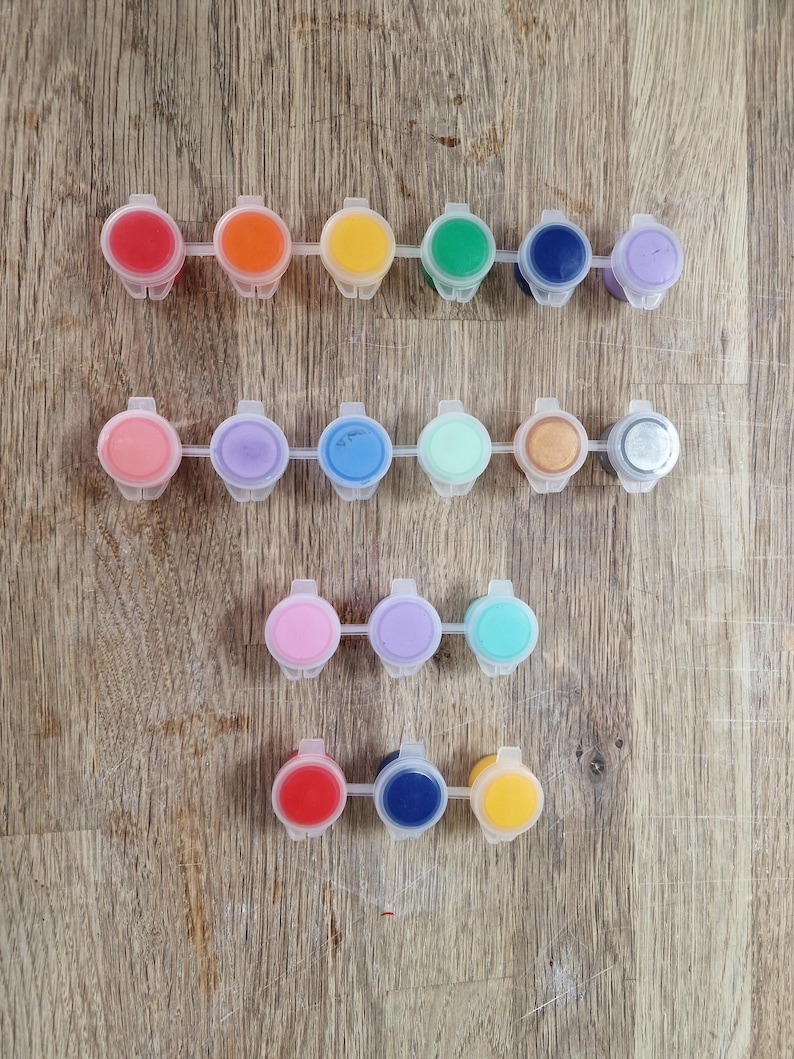 May include: A collection of small, plastic paint pots filled with various colors, including red, orange, yellow, green, blue, and purple. The paint pots are arranged in rows on a wooden surface, offering a vibrant palette for art projects.