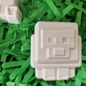 Puede incluir: Objetos blancos, cuadrados y angulosos, con un dise&ntilde;o de cara pixelada, que se asemejan a un personaje de videojuego, descansan sobre un lecho de papel triturado verde brillante. Los objetos tienen una apariencia geom&eacute;trica distinta.