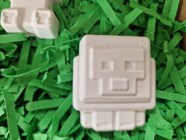 Puede incluir: Objetos blancos, cuadrados y angulosos, con un dise&ntilde;o de cara pixelada, que se asemejan a un personaje de videojuego, descansan sobre un lecho de papel triturado verde brillante. Los objetos tienen una apariencia geom&eacute;trica distinta.